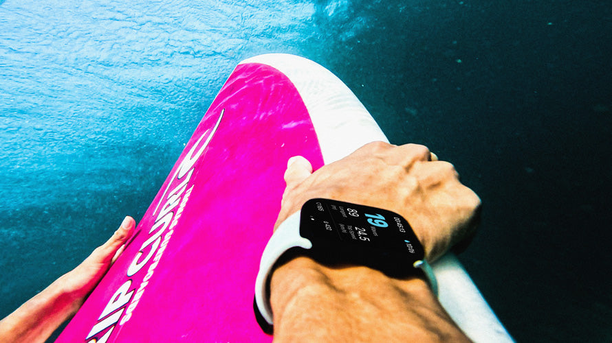 Person holding a pink and white surfboard with a digital watch on wrist, water background