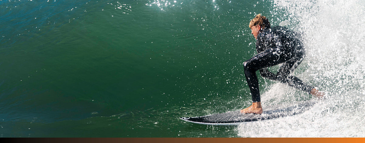 A surfer in a black wetsuit crouches on a surfboard inside a curling ocean wave with white spray, photographed outdoors in the water.