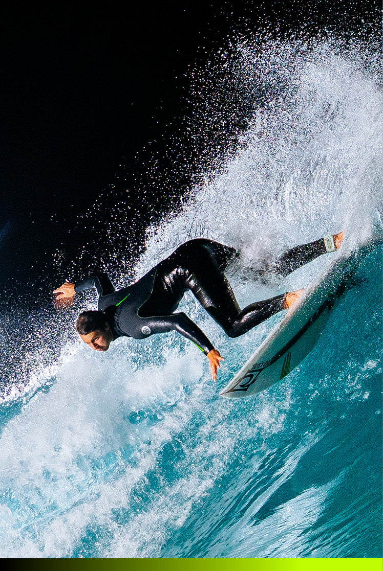 A surfer in a black wetsuit rides a white surfboard through a powerful turquoise ocean wave, with dramatic white spray and a dark sky background.