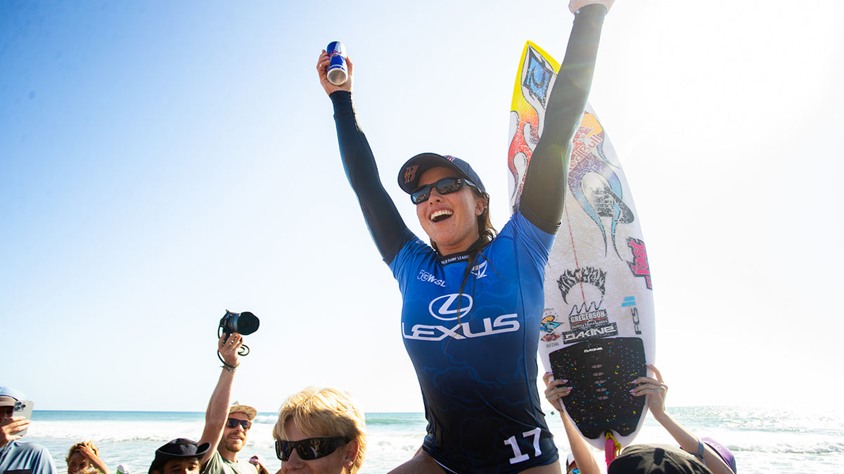 A woman in a blue and black Lexus surf jersey celebrates on a beach with her arms raised, holding a branded surfboard and a beverage can. Other beachgoers and the ocean are visible in the background.