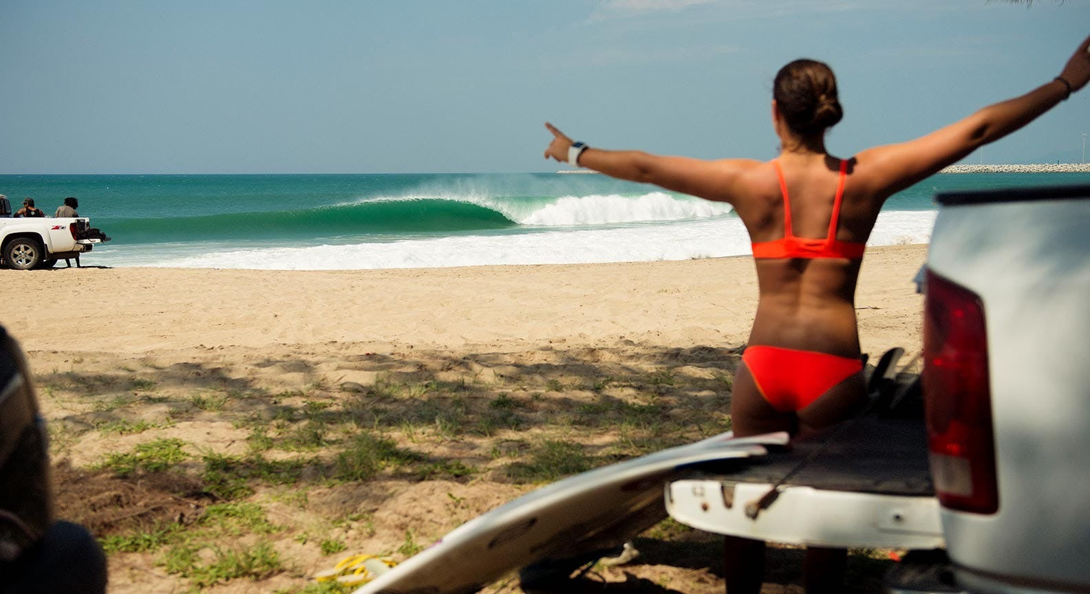 Woman in a red bikini standing on a beach with arms outstretched, near a car and surfboard.