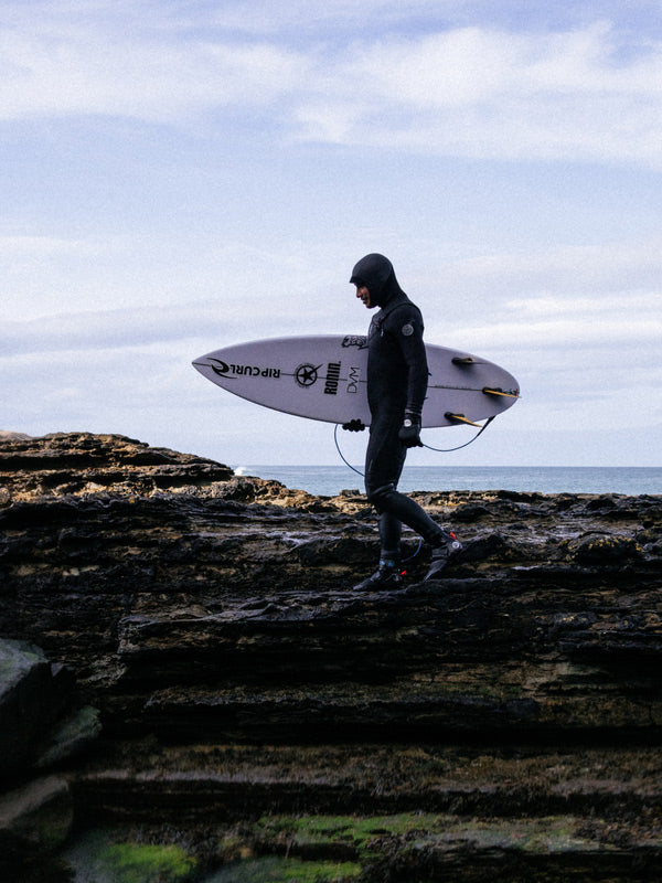 Person in a wetsuit holding a surfboard on rocky terrain with ocean and sky in the background