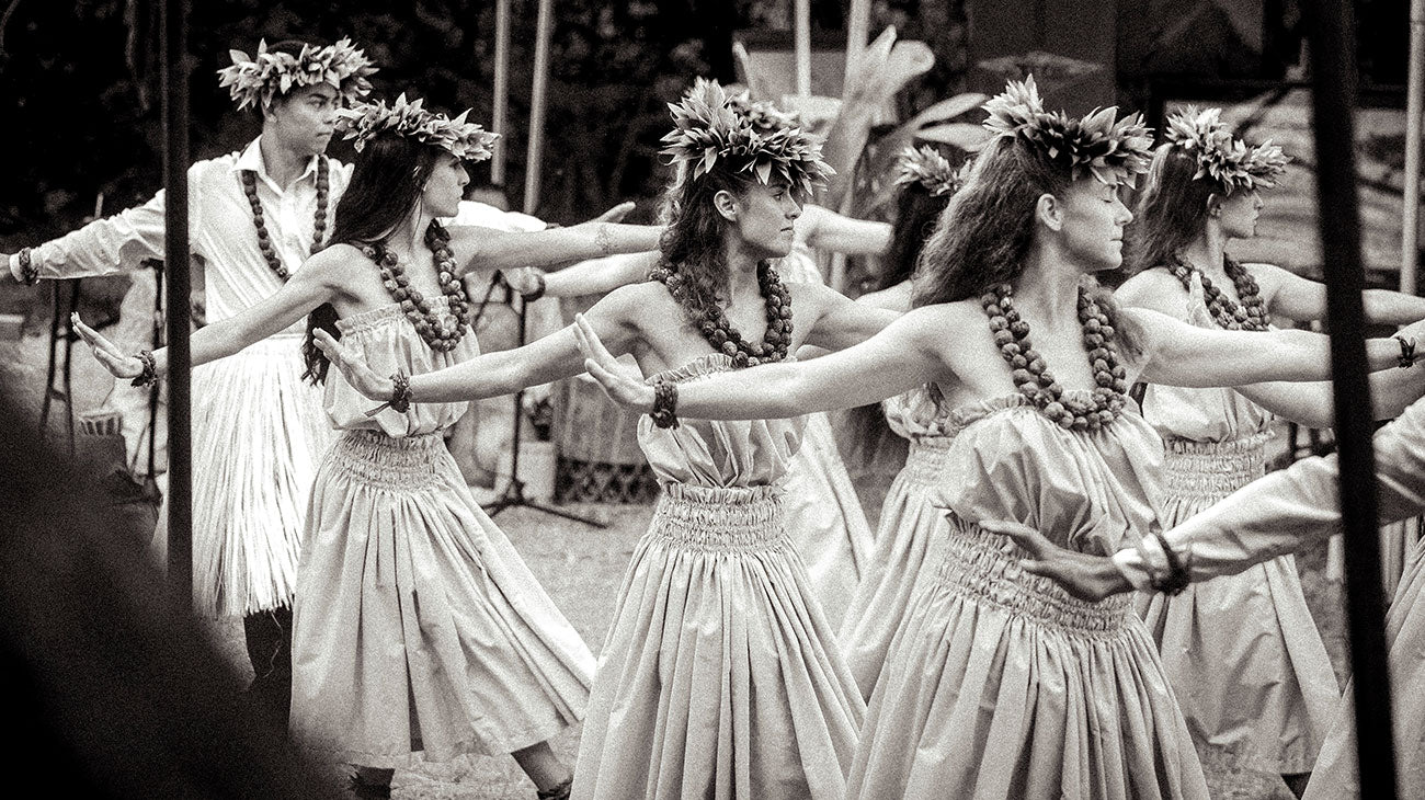 Group of dancers in traditional attire with leis and flower headbands performing a hula dance.