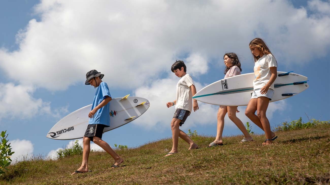 Four children walking on a grassy hill with surfboards, under a blue sky with clouds.
