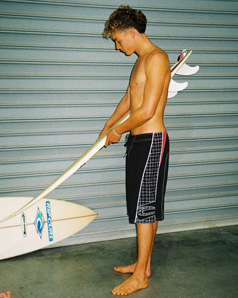 A shirtless young man stands barefoot in front of a gray metal garage door, holding a white surfboard under one arm and a paddleboard behind him. He is wearing black board shorts with white and red side stripes.