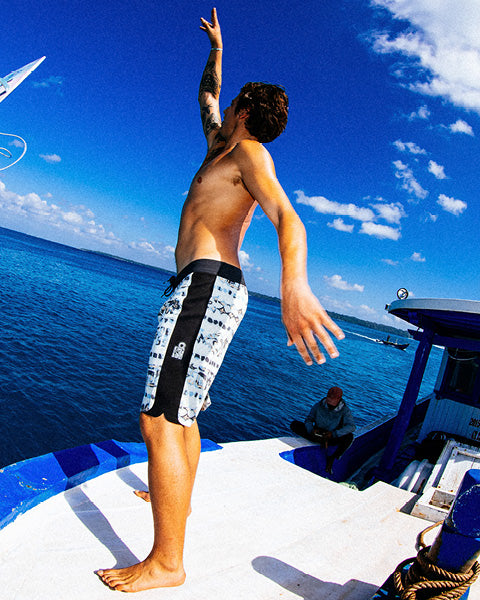 A shirtless man wearing patterned swim trunks stands on the edge of a boat, looking out over the ocean under a bright blue sky.
