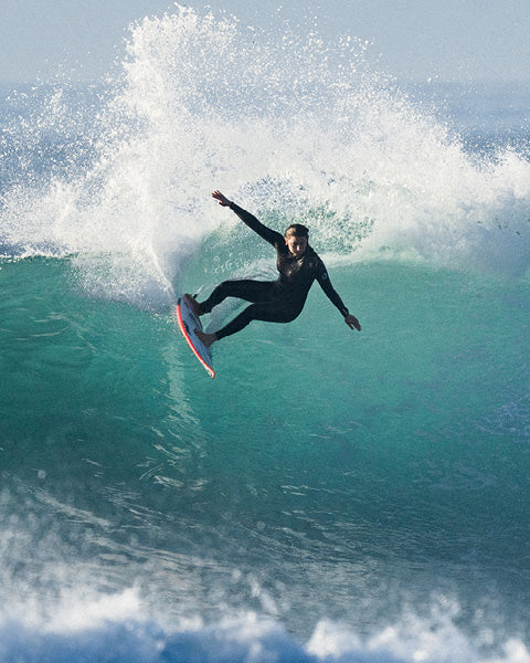 A surfer in a black wetsuit rides a large turquoise ocean wave with a dramatic spray of white water behind them.