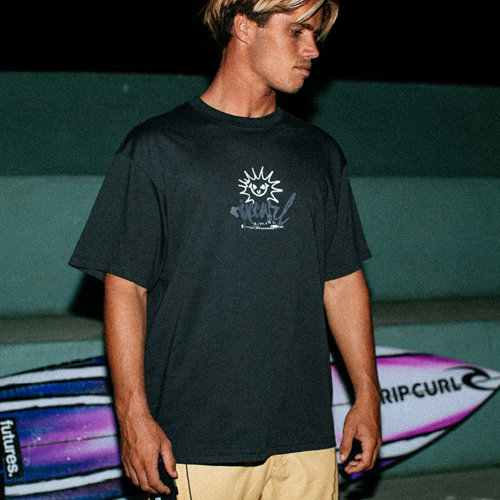 Person wearing a black t-shirt with a logo, standing in front of surfboards.