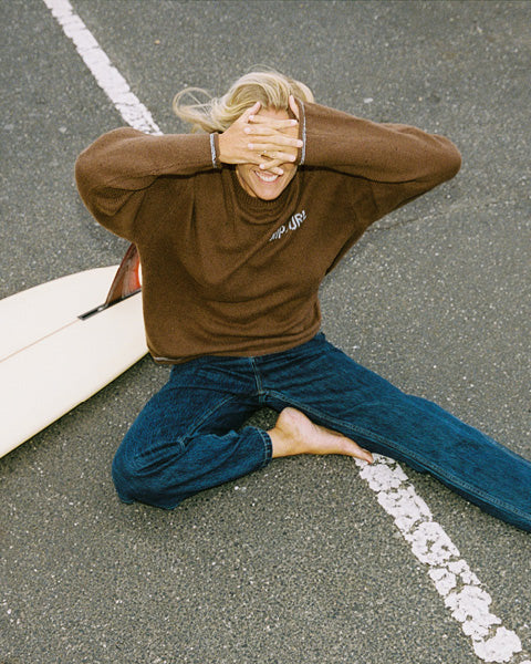 A person sitting on asphalt outdoors, wearing a brown long-sleeve turtleneck sweater with a small white 'SUN AIR' logo on the chest and blue jeans. A white surfboard is partially visible beside them.