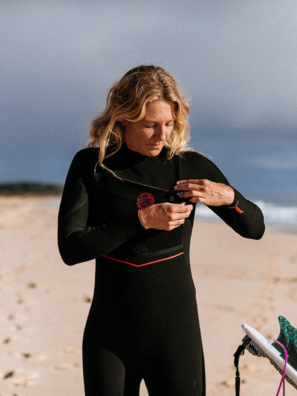 A woman wearing a black full-body wetsuit stands on a sandy beach, adjusting a strap or accessory near her chest. The wetsuit has a small red circular logo on the left chest and a thin red horizontal accent line near the waist.