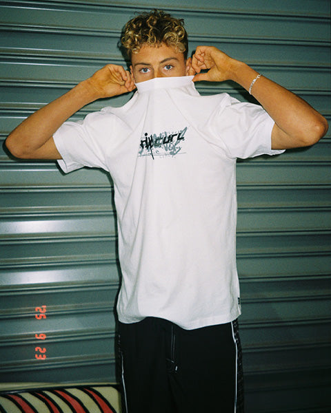 A young man wearing a white t-shirt with a black graphic print on the chest, posing with the collar pulled up to his eyes against a green metal shutter background.