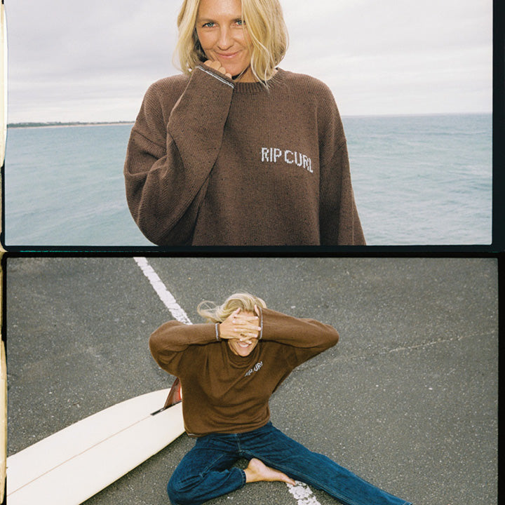 A split-screen lifestyle image of a woman wearing a brown ribbed knit sweater with 'RIP CURL' embroidered in white on the chest. The top panel shows her standing by the ocean, and the bottom panel shows her sitting on a surfboard on a paved street.