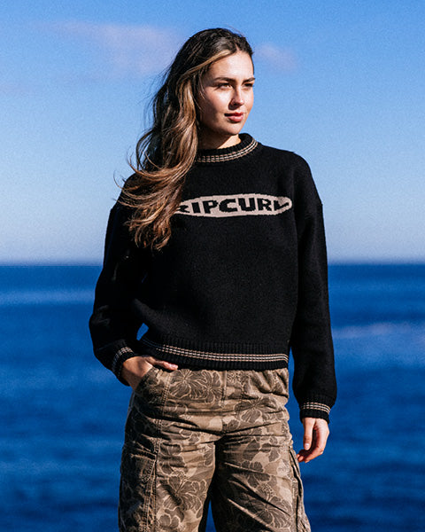Person wearing a black sweater with 'Epifani' branding against a blue sky and ocean background