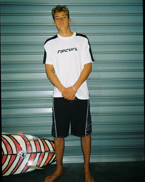 Person wearing a Rip Curl shirt standing next to a surfboard against a metallic shutter background