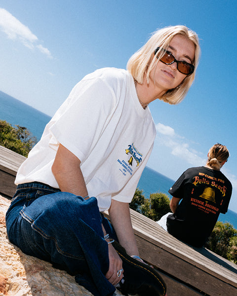 Person wearing a white t-shirt with a logo, sitting on a ledge with a scenic background.