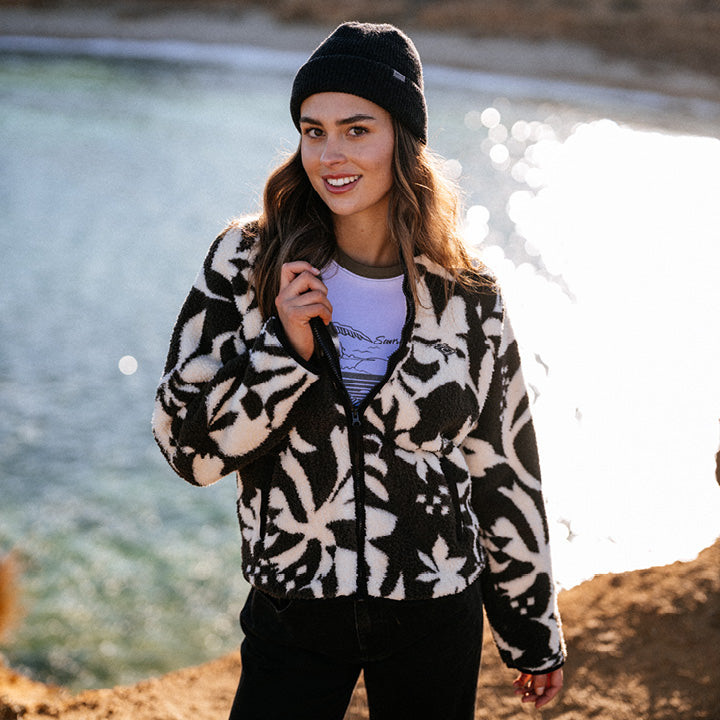 Woman wearing a patterned jacket and beanie standing by a body of water.