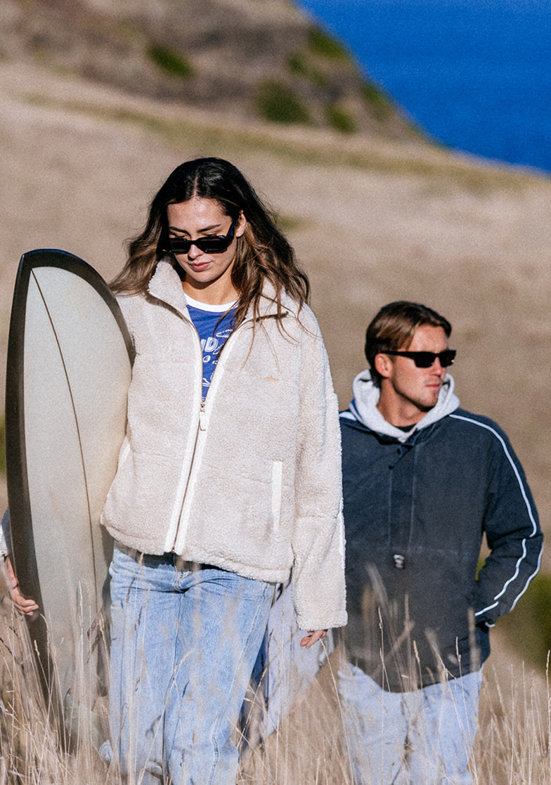 Two people walking on a beach with a surfboard, wearing sunglasses and casual clothing.