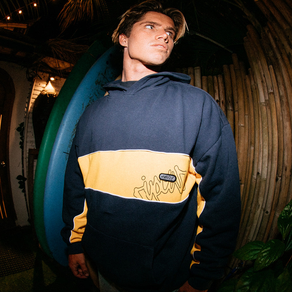 Person wearing a navy and yellow hoodie with a logo, standing in a tropical setting.