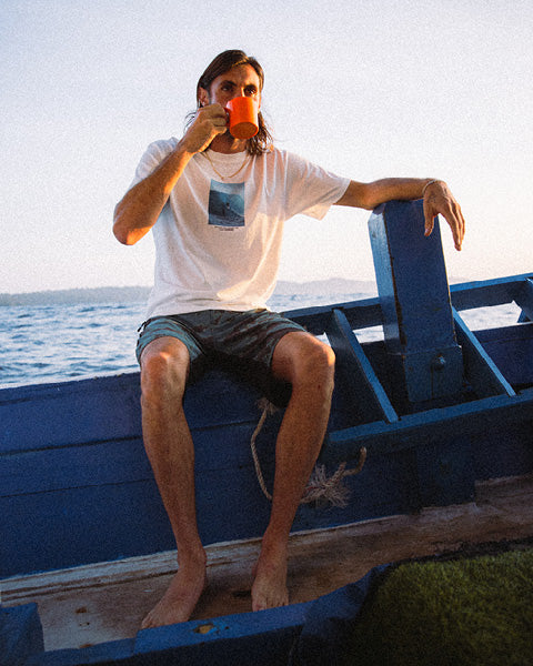 A man with long dark hair sits barefoot on the edge of a blue boat, drinking from an orange mug. He is wearing a white graphic t-shirt and dark shorts, with water and a pale sky in the background.