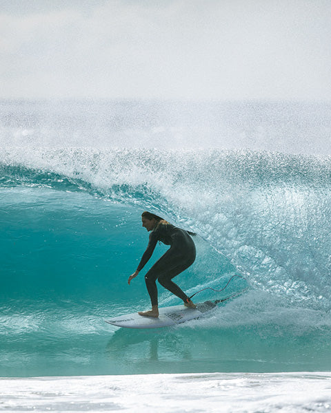 A surfer in a black wetsuit rides a turquoise ocean wave on a white surfboard, with white spray and a hazy sky in the background.