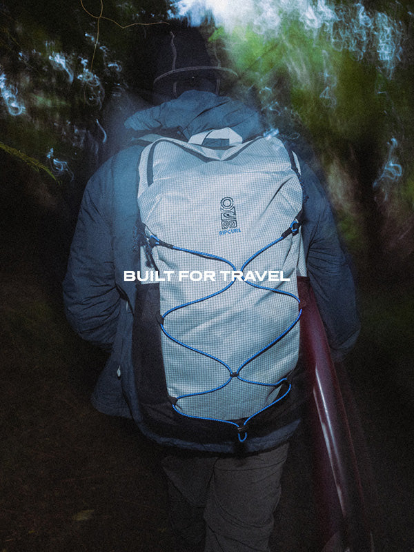 A person wearing a light gray and black backpack with blue compression straps, photographed from behind in a dark outdoor setting. The backpack has a visible logo and the text 'BUILT FOR TRAVEL' overlaid across the center.