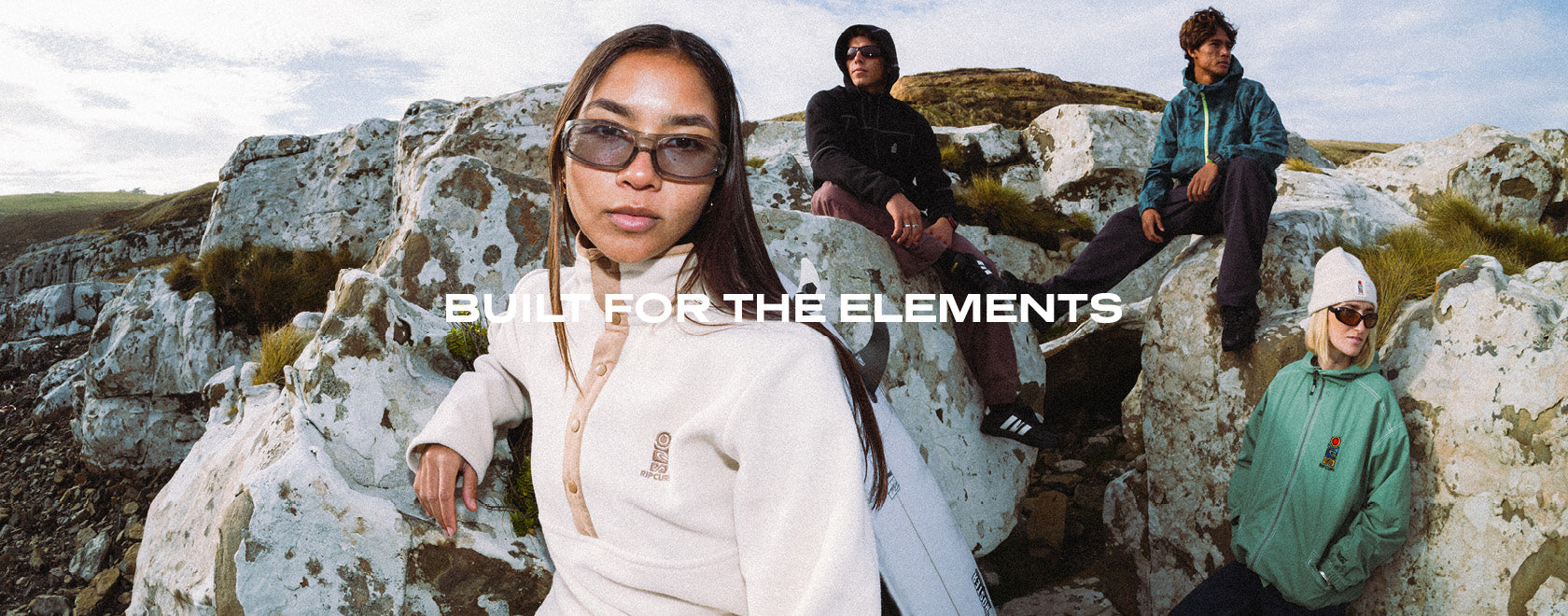 A group of four people wearing outdoor apparel and sunglasses posing on large white rocks in a rugged landscape under a cloudy sky. The text 'BUILT FOR THE ELEMENTS' is overlaid across the center.