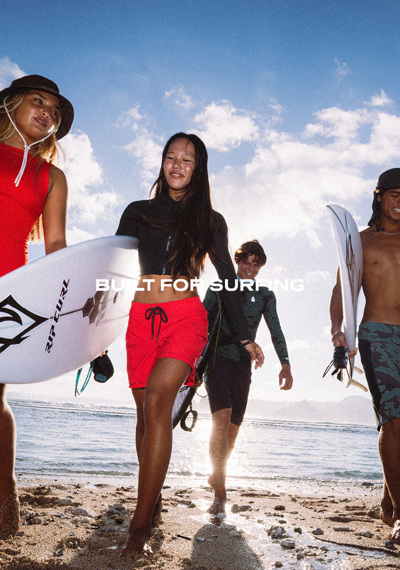 A group of surfers walking along a sandy beach under a blue sky, with one central woman in a black top and bright red shorts carrying a surfboard, and other surfers around her carrying boards. Large white text across the center reads 'BUILT FOR SURFING'.