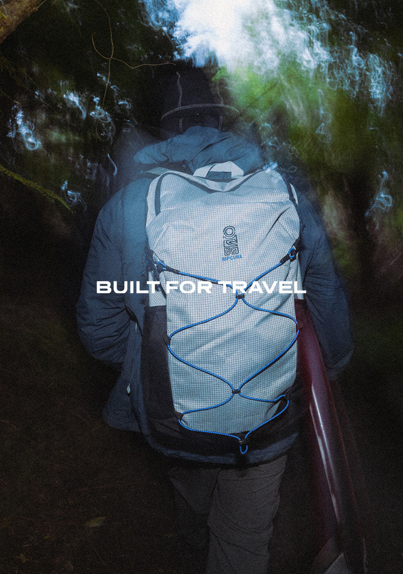 A person wearing a light gray and black travel backpack with blue compression straps, seen from behind while walking through a dark, misty outdoor environment. The text 'BUILT FOR TRAVEL' is overlaid across the center.