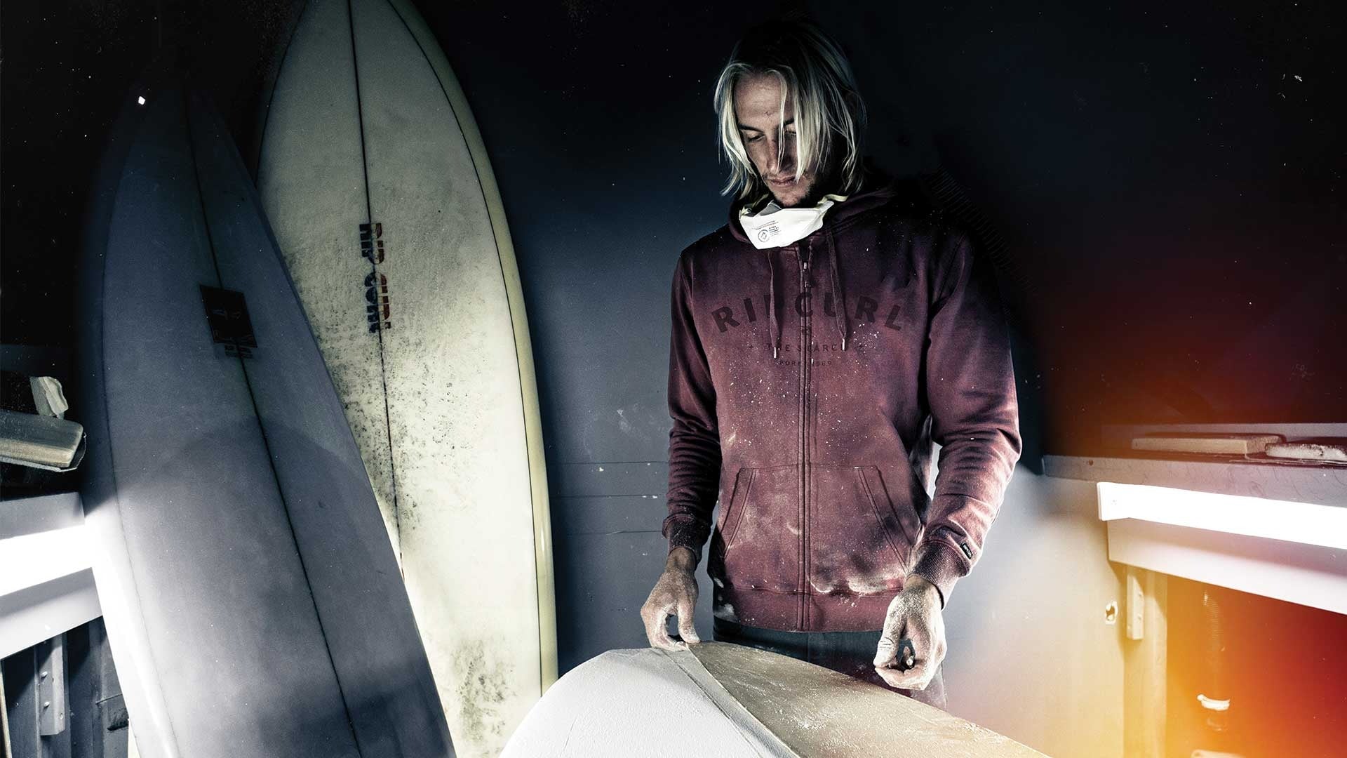 Person working on a surfboard in a workshop with surfboards in the background